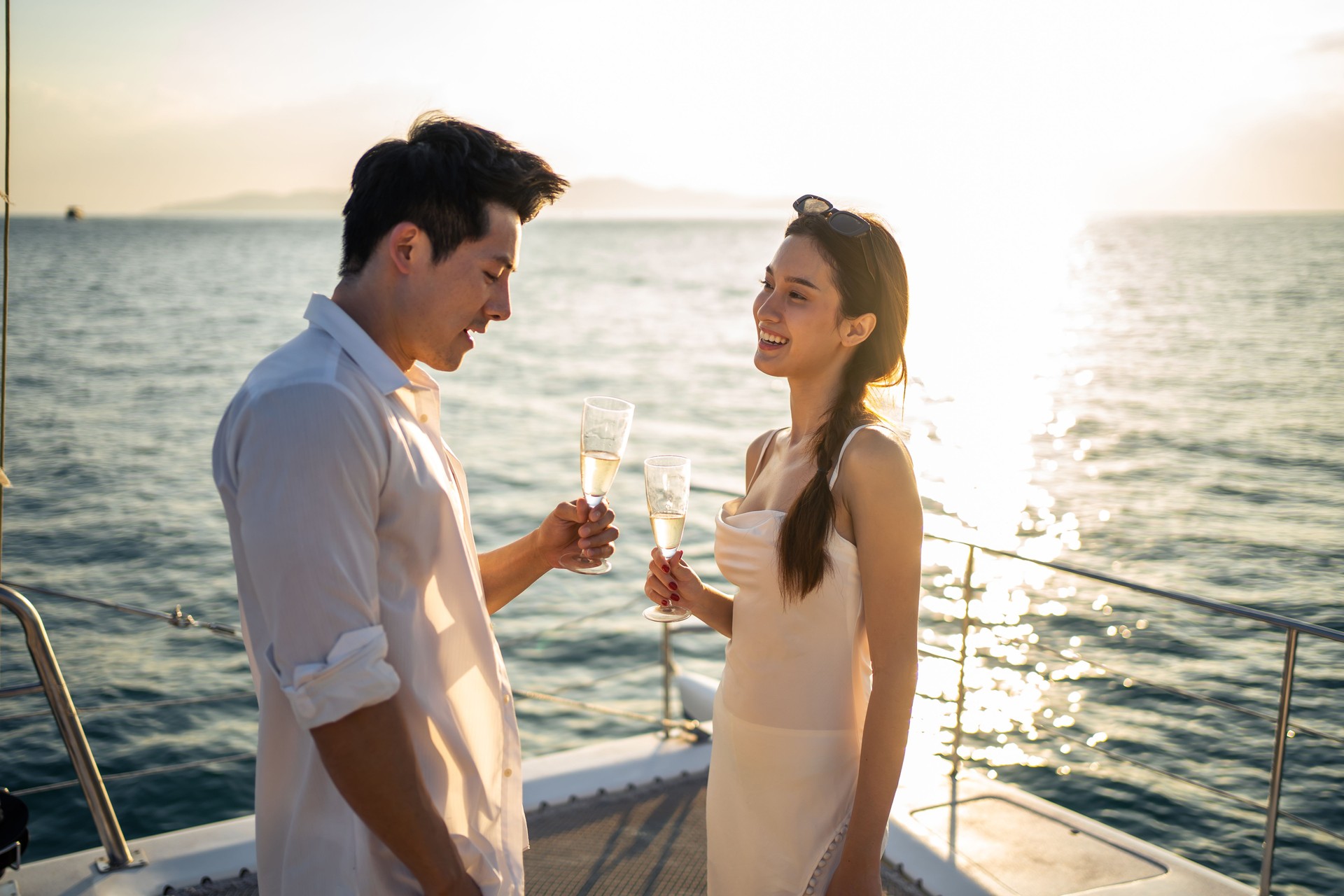 Asian beautiful couple drinking champagne while having party in yacht. Attractive man and woman hanging out, celebrating anniversary honeymoon trip while catamaran boat sailing during summer sunset.