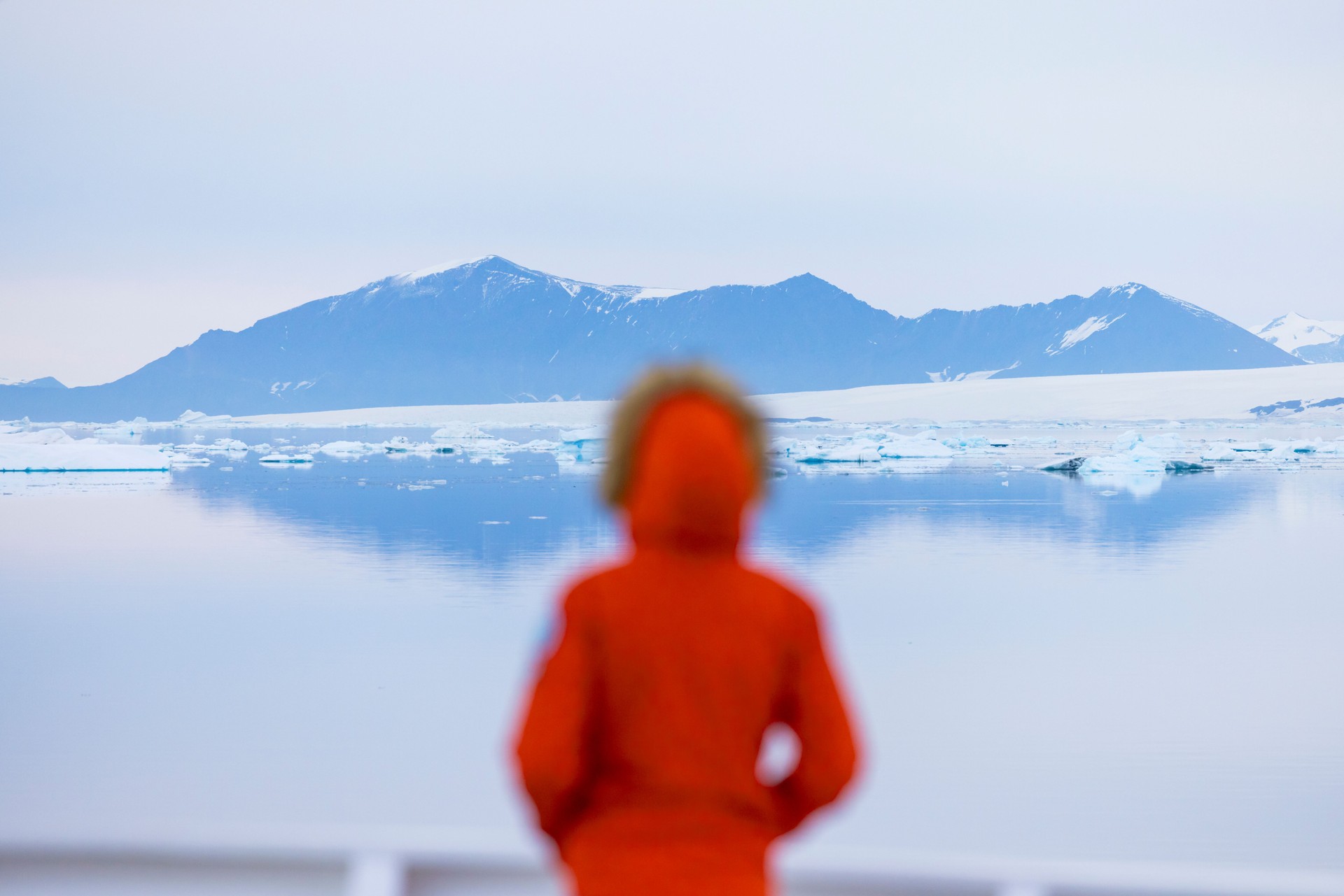 A woman wearing a warm red jacket is standing on the bow of a ship in Antarctica.